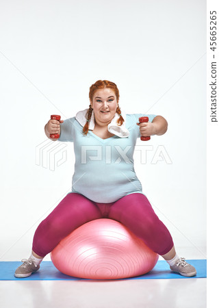 Red haired, chubby woman is sitting on the ball and keep the dumbbells Red haired, chubby woman is sitting on the ball and keep the dumbbells 45665265