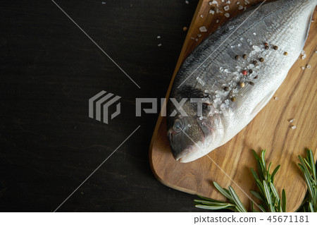 top view of fresh raw dorada fish with rosemary, pepper and salt on a wooden board and a black table top view of fresh raw dorada fish with rosemary, pepper and salt on a wooden board and a black table 45671181