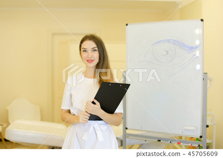 Young cosmetologist standing with black folder near placard with drawn eye at cosmetology cabinet. 45705609