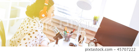 Beautiful young girl sits in headphones and with a notepad at the table in the office and speaks 45707222