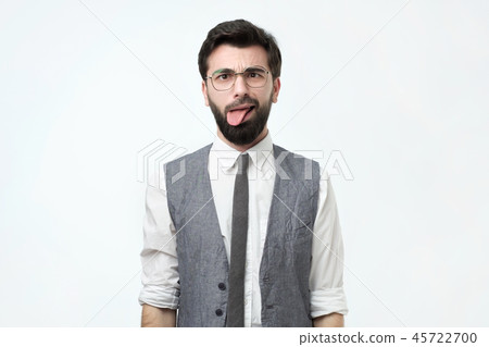 Unhappy irritated bearded young man wears white shirt and vest feels displeased, shows tongue Unhappy irritated bearded young man wears white shirt and vest feels displeased, shows tongue 45722700