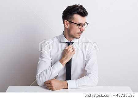 Portrait of a young man with white shirt sitting at the table in a studio. Portrait of a young man with white shirt sitting at the table in a studio. 45725153