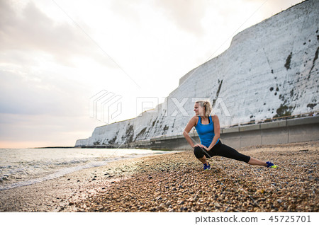 Young sporty woman runner in blue sportswear stretching on the beach outside. 45725701