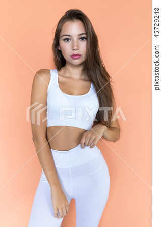 Attractive young woman in white sports top and leggings on a beige background. Attractive young woman in white sports top and leggings on a beige background. 45729248