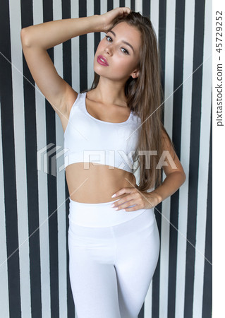 Attractive young woman in white sports top and leggings on striped background. Attractive young woman in white sports top and leggings on striped background. 45729252