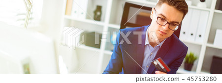 A young man is sitting at a table in the office, holding a bank card in his hand and typing on a A young man is sitting at a table in the office, holding a bank card in his hand and typing on a 45753280