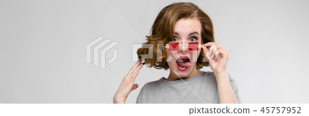 Charming young girl in a gray T-shirt on a gray background. Girl in red square-eyed glasses. 45757952