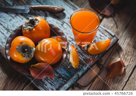 Ripe orange persimmon fruit and persimmon leaves in a brown plate on a brown wooden table Ripe orange persimmon fruit and persimmon leaves in a brown plate on a brown wooden table 45766726
