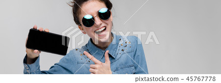 Young brunette girl in round glasses. Hairs are gathered in a bun. Girl with a black phone. 45767778