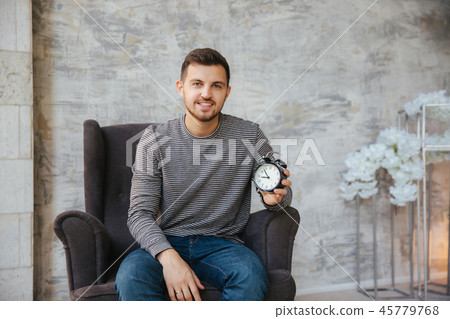 A young man sits on a chair and hold a clock in his hands. Time to stant. Handsome man smile and A young man sits on a chair and hold a clock in his hands. Time to stant. Handsome man smile and 45779768