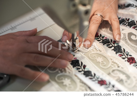 Seamstress hands connecting white fabric embroidered details with stripes of lace on sewing machine. Seamstress hands connecting white fabric embroidered details with stripes of lace on sewing machine. 45786244