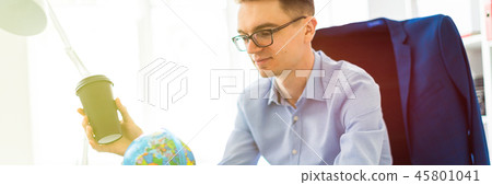 A young man sits in the office at a computer desk and holds a glass of coffee in his hand. A young A young man sits in the office at a computer desk and holds a glass of coffee in his hand. A young 45801041