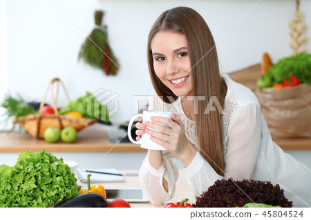 Young happy woman holding white cup and looking at the camera while sitting at wooden table in the Young happy woman holding white cup and looking at the camera while sitting at wooden table in the 45804524