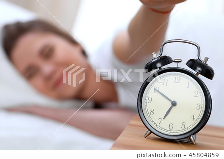 Alarm clock opposite of sleepy young woman stretching hand to ringing alarm willing turn it off Alarm clock opposite of sleepy young woman stretching hand to ringing alarm willing turn it off 45804859