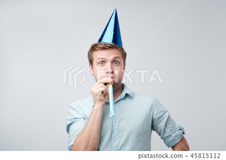 cheerful young man having fun on party wearing blue denim shirt and holiday hat, blowing party horn. cheerful young man having fun on party wearing blue denim shirt and holiday hat, blowing party horn. 45815112
