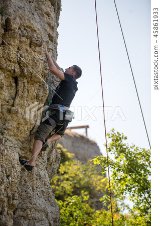 Photo of athlete man in helmet clambering over rock against background of green trees Photo of athlete man in helmet clambering over rock against background of green trees 45861933
