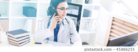A beautiful young girl in a white robe is sitting at the table, talking on the phone and holding her 45866255