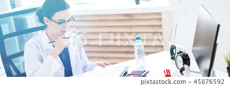A beautiful young girl in a white robe is sitting at a computer desk with documents and a pen in her 45876592
