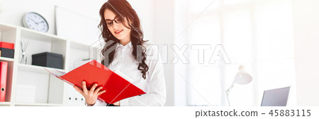 Beautiful young girl stands in office, leaning on desk and scrolls through red folder with documents Beautiful young girl stands in office, leaning on desk and scrolls through red folder with documents 45883115