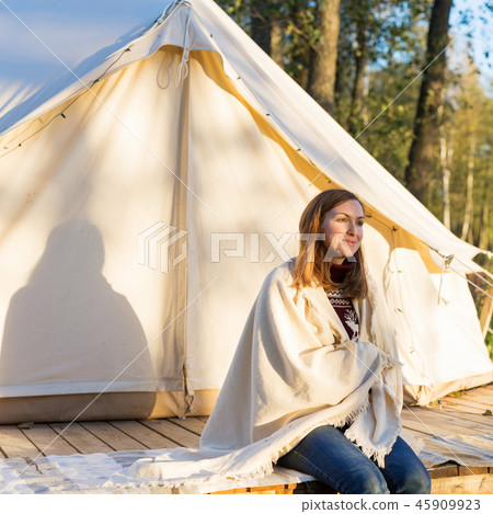 Young woman wraps a blanket over herself while sitting near canvas tent Young woman wraps a blanket over herself while sitting near canvas tent 45909923