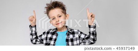 A handsome boy in a plaid shirt, blue shirt and jeans stands on a gray background. Boy smiling and A handsome boy in a plaid shirt, blue shirt and jeans stands on a gray background. Boy smiling and 45937149