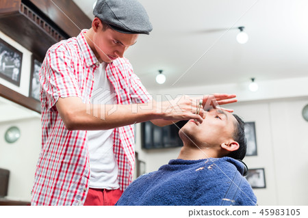 Male barber wearing baseball cap shaving with razor 45983105