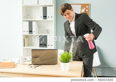 Business people, fun and potted plants concept - young funny businessman watering plant at office Business people, fun and potted plants concept - young funny businessman watering plant at office 46003800