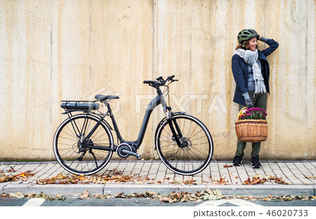 Active senior woman with electrobike standing outdoors in town, leaning against bike. Active senior woman with electrobike standing outdoors in town, leaning against bike. 46020733