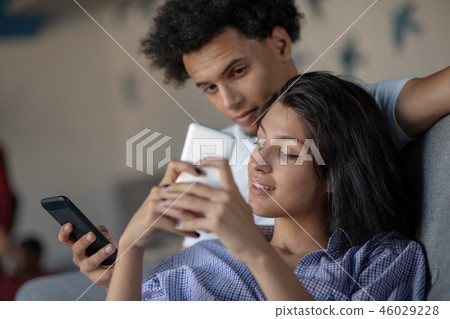 Attractive couple sitting on couch together looking at smartphone at home in the living room Attractive couple sitting on couch together looking at smartphone at home in the living room 46029228