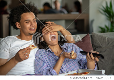 Happy couple watching tv while eating pizza. Shallow depth of field, focus on the man Happy couple watching tv while eating pizza. Shallow depth of field, focus on the man 46030007