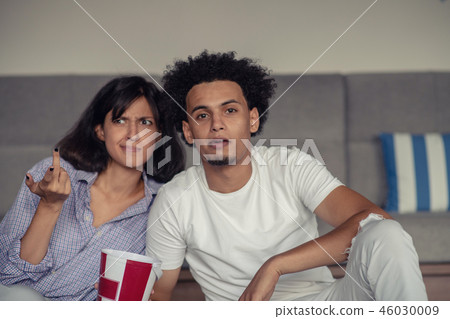 Image of smiling couple sitting on floor near sofa at home and looking aside while eating popcorn 46030009