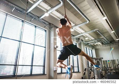Fit young topless man in gym climbing a rope. Fit young topless man in gym climbing a rope. 46046502