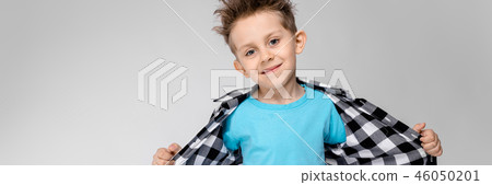 A handsome boy in a plaid shirt, blue shirt and jeans stands on a gray background. The boy pulls his 46050201