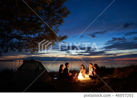 Night summer camping on shore. Group of young tourists around campfire near tent under evening sky Night summer camping on shore. Group of young tourists around campfire near tent under evening sky 46057201