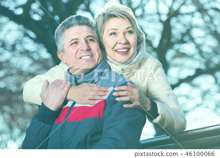 positive mature married couple sitting on park bench 46100066