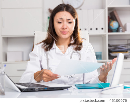 Upset girl doctor in uniform with papers at laptop in hospital Upset girl doctor in uniform with papers at laptop in hospital 46100976