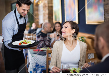 Waiter with dishes serving man and woman friendly company indoors Waiter with dishes serving man and woman friendly company indoors 46101756