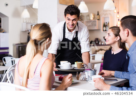 Polite happy waiter bringing ordered dishes to friends in tearoom 46104534