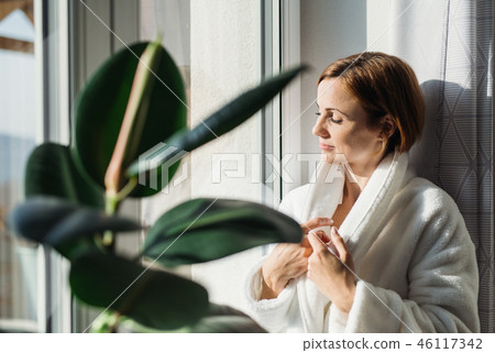 A young woman with bath robe standing indoors by a window in the morning. 46117342