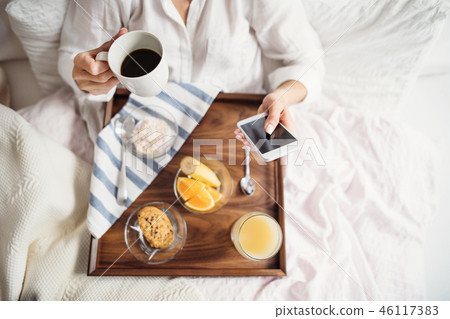 A midsection of woman with breakfast in bed in the morning, using smartphone. A top view. A midsection of woman with breakfast in bed in the morning, using smartphone. A top view. 46117383
