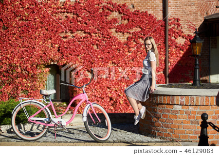 Attractive blond girl at pink lady bicycle on sunny day on wall overgrown with red ivy background. 46129083