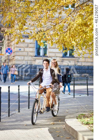 Young tourist couple, handsome man and pretty blond woman riding tandem bicycle along city street. 46129087