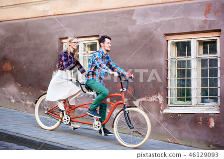 Young tourist couple, handsome man and pretty blond woman riding tandem bicycle along city street. 46129093