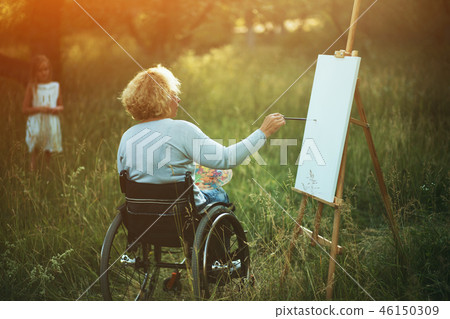 Woman in wheelchair drawing on easel outside Woman in wheelchair drawing on easel outside 46150309