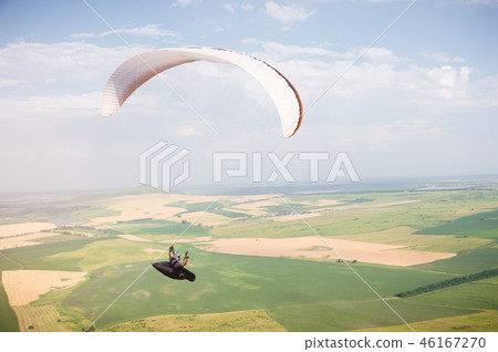 Professional paraglider in a cocoon suit flies high above the ground against the sky and fields 46167270