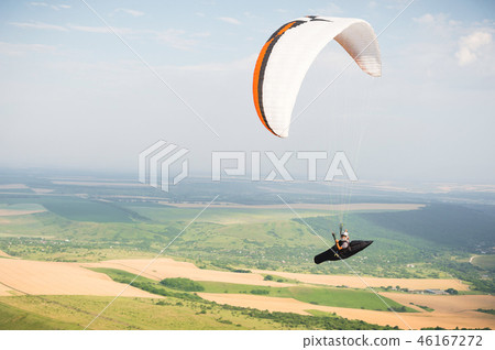 Professional paraglider in a cocoon suit flies high above the ground against the sky and fields 46167272