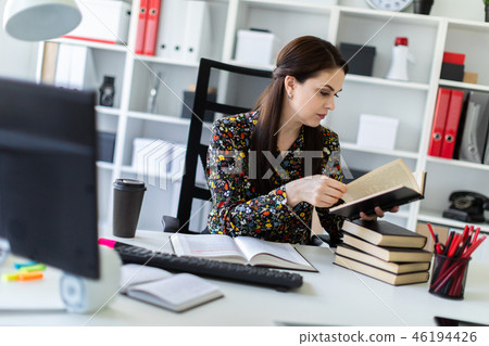 A young girl sitting in the office at the computer Desk and working with a book. A young girl sitting in the office at the computer Desk and working with a book. 46194426