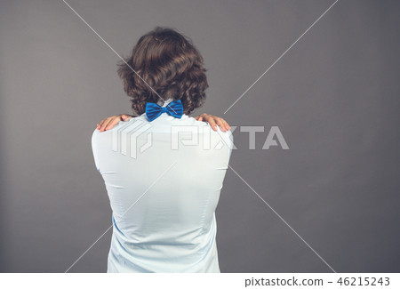 Young man in blue shirt and bow tie hugs himself on grey background. Rear view. His hands lie on the 46215243