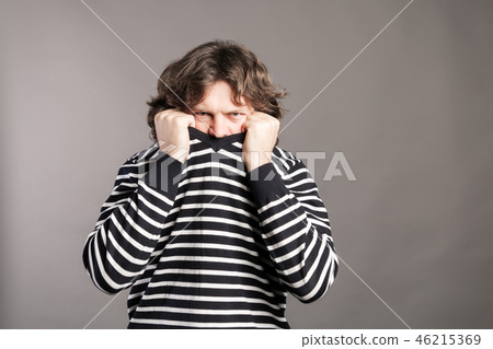 Young man pulling black and white striped sweater over head against gray background. Angry male 46215369