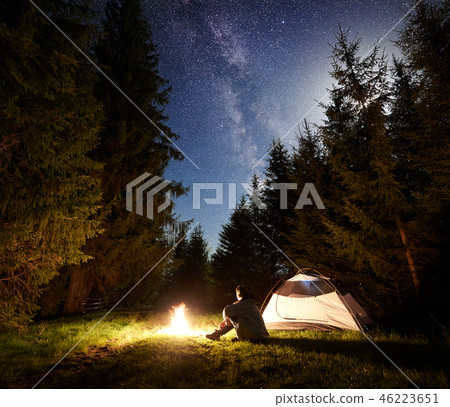 Male hiker enjoyng night camping near tourist tent at campfire under blue starry sky and Milky way Male hiker enjoyng night camping near tourist tent at campfire under blue starry sky and Milky way 46223651